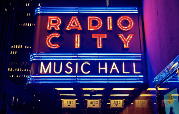 Das erleuchtete Radio City Music Hall in New York City mit hohen Gebäuden drumherum, mit einer 'Radio City'-Leuchtschrift und zusätzlichen Schildern auf der rechten Seite.