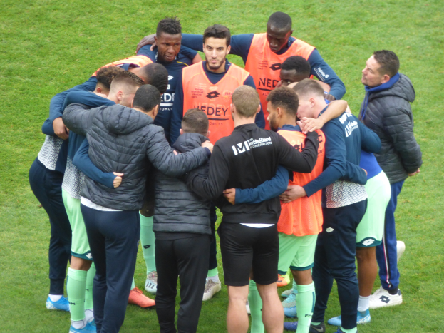 A group of men in jackets huddle together in a circle on a soccer field.