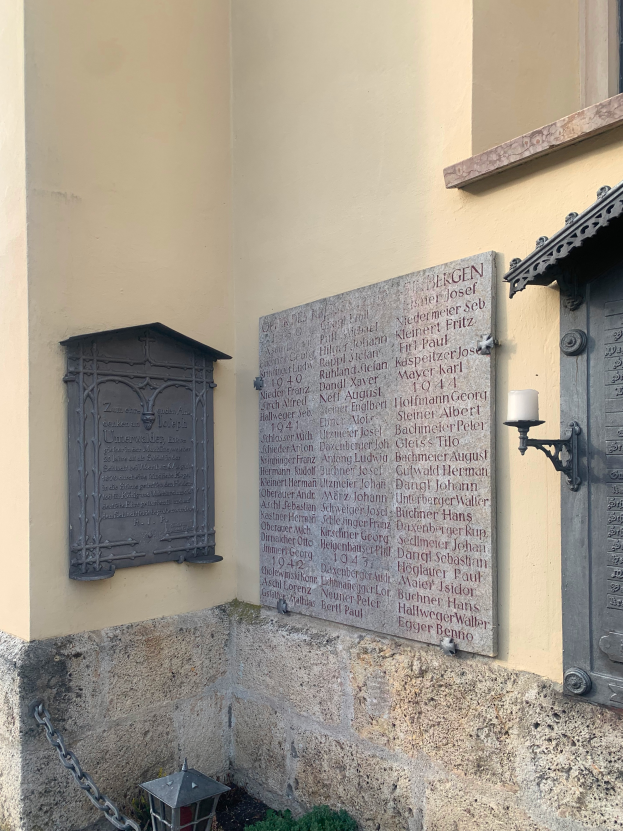 Ein Gebäude mit einer Gedenktafel, einem Fenster, einer Tür, einer Lampe, einer Kette und einigen Pflanzen, das Holocaust-Opfern und Kriegsopfern gedenkt.