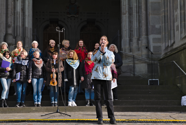 Eine Gruppe von Menschen, einige mit Musikinstrumenten, steht vor einem Gebäude mit Treppen und Geländern, während eine Frau in der Mitte in ein Mikrofon singt, umgeben von zusätzlichen Mikrofonen auf Ständern.