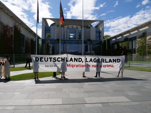 Eine Gruppe von Menschen hält eine Fahne mit der Aufschrift "Deutschland, Lagerland Migration ist kein Verbrechen" vor einem Gebäude mit Fenstern, umgeben von Gras, einem Metallzaun, Pflanzen, Bäumen und Fahnenstangen unter einem bewölkten Himmel.