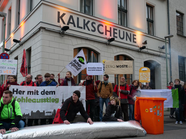 Eine Gruppe von Menschen steht vor einem Gebäude mit Schildern und Plakaten, zwei Personen sitzen im Vordergrund und ein Müllcontainer rechts, während einer Demonstration in Deutschland.