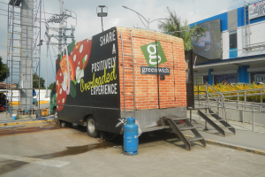 Lastwagen vor einem Gebäude mit einer Tafel, auf der "Teilen Sie eine positiv überladene Erfahrung" steht, mit Stufen, einem Zylinder auf dem Boden, Geländern, Strommasten mit Drähten, Laternenmasten, Bäumen und einem bewölkten Himmel.