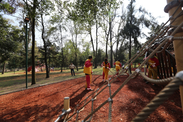 Gruppe von Menschen, die einen Seilparcours in einem Park mit Bäumen im Hintergrund und bodenbedecktem Boden absolvieren, mit einem Pfahl mit Seilen auf der rechten Seite.