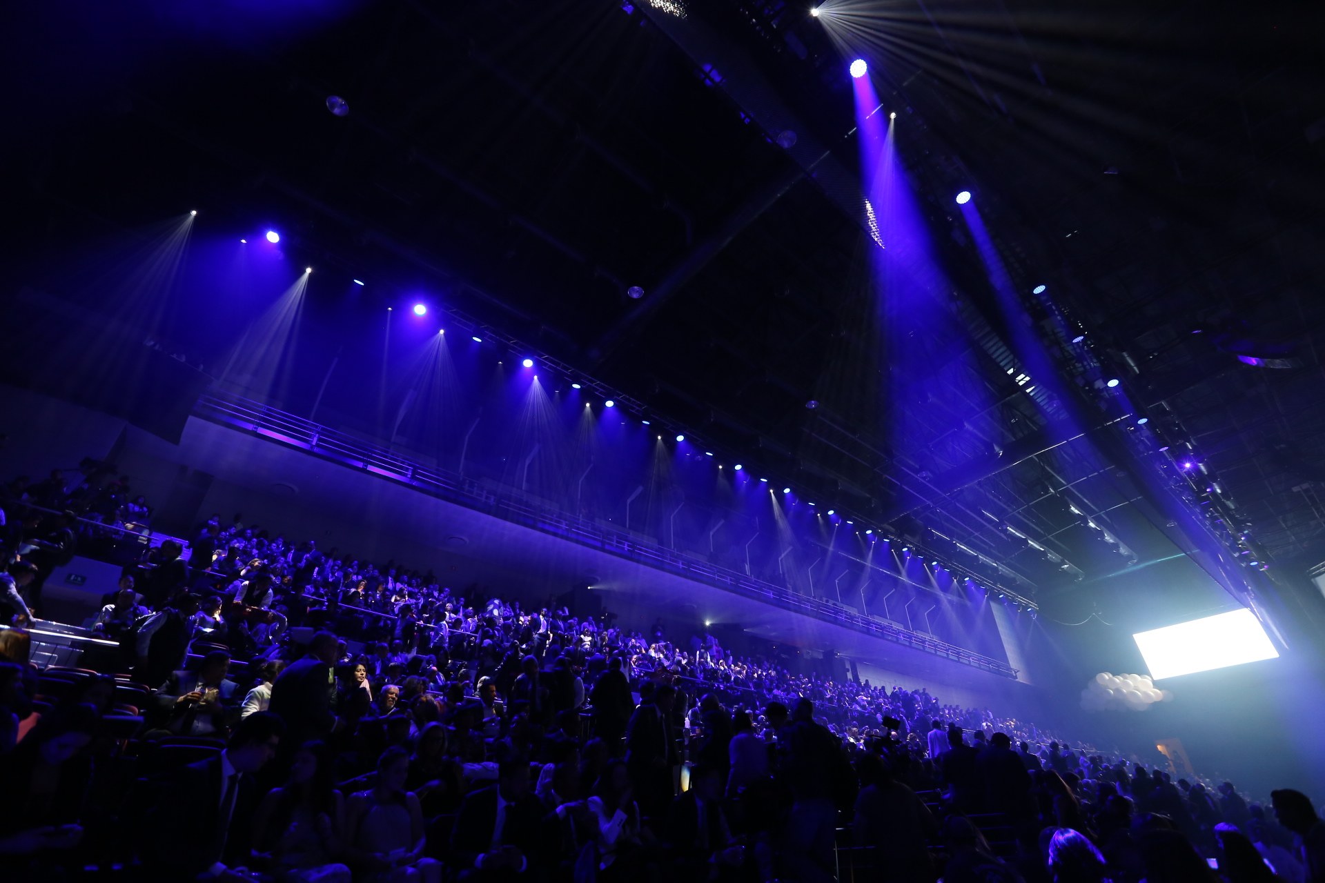 Eine große Menschenmenge sitzt in einem abgedunkelten Konzertsaal, der einer Bühne mit Deckenleuchten und einem Bildschirm auf der rechten Seite gegenüberliegt.
