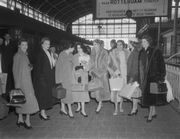 Schwarzes und weißes Bild einer Gruppe von Frauen, die zusammen auf einem Bahnhof stehen, jede hält eine Tasche, mit einem Zug auf der linken Seite und Geländern und einer Tafel auf der rechten Seite.