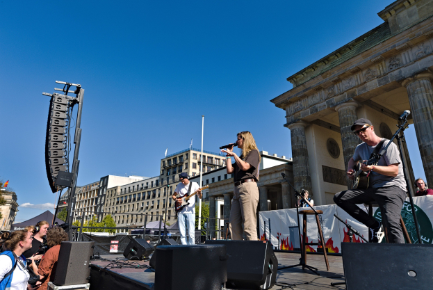 Eine Gruppe von Menschen, die auf einer Bühne Musik spielen, mit dem Brandenburger Tor im Hintergrund, begleitet von Lautsprechern und Bühnenausrüstung unter einem klaren blauen Himmel.