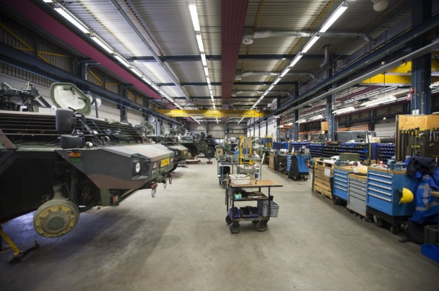 Ein großes Lager mit Maschinen, Fahrzeugen auf der linken Seite, Wagen mit Gegenständen, Holzboxen und Deckenbeleuchtung.