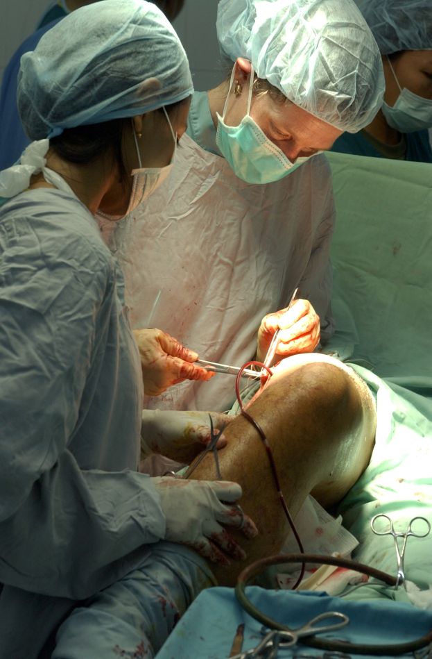 Eine Gruppe von Chirurgen in Masken, Hauben und Handschuhen operiert einen Patienten auf einem Bett, umgeben von medizinischen Instrumenten und Materialien in einem Operationssaal.