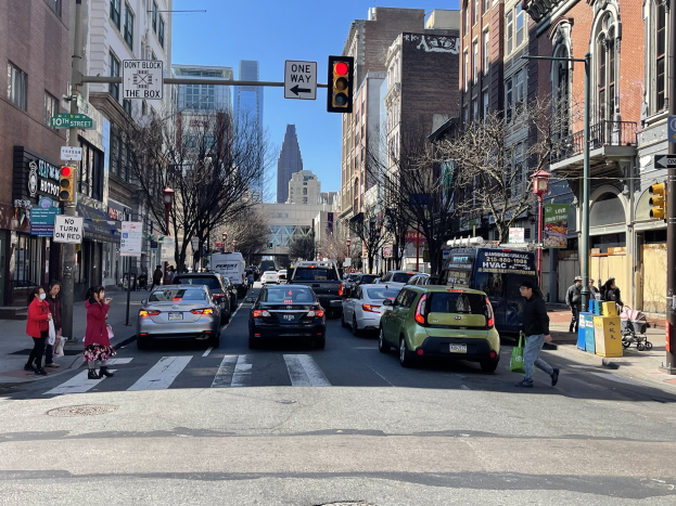 Eine belebte Stadtstraße mit Fahrzeugen, Fußgängern, Bäumen, Gebäuden, Schildern, Laternen, einer Ampel und einem geparkten Van unter einem sichtbaren Himmel.