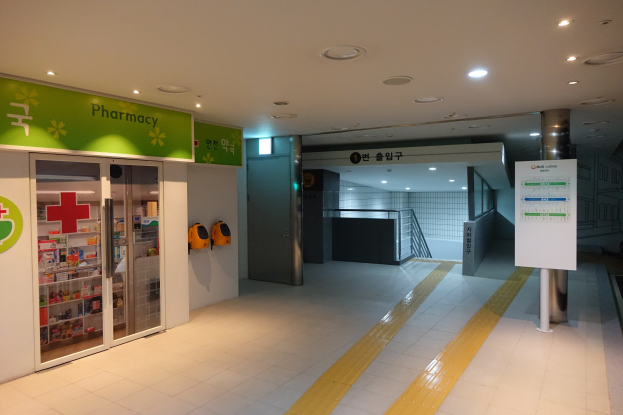 Apothekeneingang mit einem "Apotheke"-Schild auf der linken Seite, einer Tafel mit Text auf der rechten Seite, beleuchteten Lampen darüber und Hintergrundobjekten.