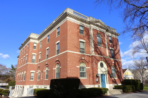 Ein großes Backsteingebäude mit einer blauen Tür, umgeben von Bäumen und Pflanzen, mit einer Straße im Vordergrund und einem bewölkten Himmel im Hintergrund, das die neue Campus der University of Pennsylvania darstellt.