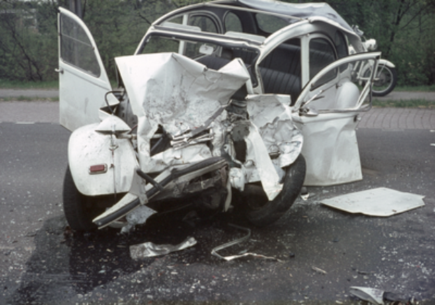 Ein stark beschädigtes Auto mit schwerer Frontschäden am Straßenrand, mit Bäumen, einem Gebäude und einem anderen Fahrzeug im Hintergrund.