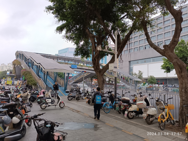 Eine belebte Straße mit parkenden E-Scootern, Passanten auf dem Gehweg, Bäumen, Gebäuden und einem bewölktem Himmel mit einem Wasserzeichen.