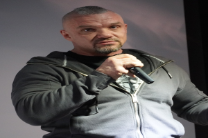 Ein Mann in einer schwarzen Kapuzenjacke und einem schwarzen Anzug mit einem weißen Hemd, der ein Mikrofon hält und selbstbewusst vor einer Wand mit zurückgekämmtem Haar steht.