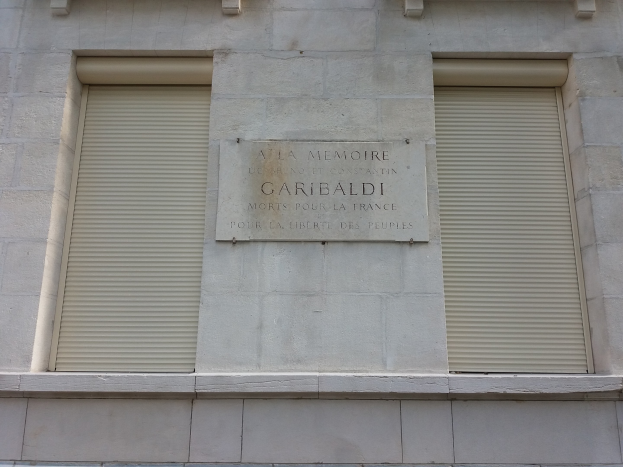 Zweistöckiges Gebäude mit zwei Fenstern mit Läden und einer Tafel an der Außenwand, wahrscheinlich ein Denkmal.