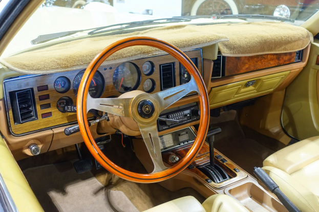 Innenansicht eines Autos mit Lenkrad, Armaturenbrett, Tacho, Autoanzeige, Ganghebel, Sitzen und Scheibenwischer, wobei andere Fahrzeuge durch die Windschutzscheibe sichtbar sind.