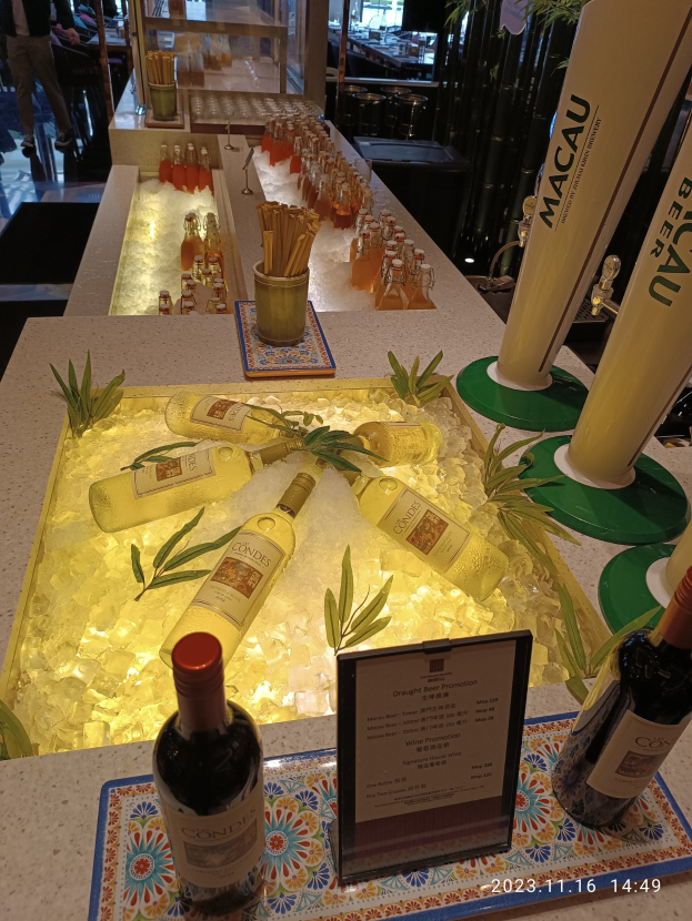 Ein Tisch mit Weinflaschen und Eiswürfeln, umgeben von Pflanzen und anderen Gegenständen, in einer festlichen Outdoor-Atmosphäre mit Menschen, Möbeln und Glastüren beim Macau-Bierfest.