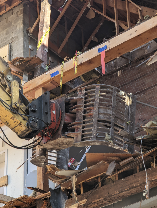 Ein Bagger arbeitet an der Seite eines teilweise abgerissenen Gebäudes, umgeben von Holzplanken und Schutt.
