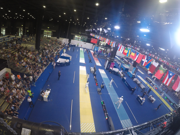 Großer Hallenstadium mit Zuschauern bei einer Turnwettbewerb, auf beiden Seiten sitzend und auf dem Boden stehend, unter Deckenleuchten mit Flaggen im Hintergrund.