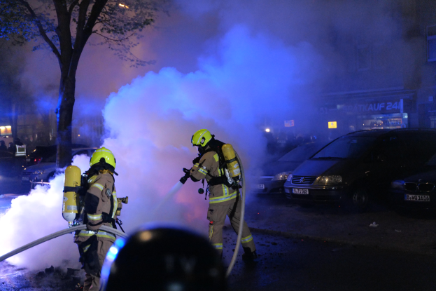 Feuerwehrleute mit Helmen und Schläuchen vor einem brennenden Auto mit Rauchwolken stehen, mit Bäumen, Gebäuden und anderen Gegenständen im Hintergrund.