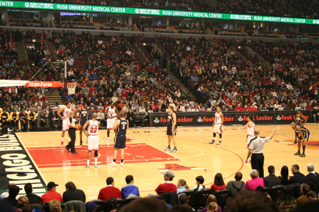 Eine Gruppe von Menschen in einem Stadion, einige halten einen Basketball, andere schauen von der Zuschauertribüne zu, und sichtbare Werbetafeln um den Basketballplatz herum.