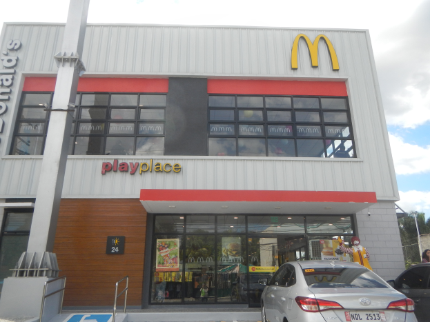 Ein McDonald's-Restaurant mit parkenden Autos davor, umgeben von Bäumen und Polen unter einem klaren blauen Himmel, mit Glaswänden, Fenstern und einem "McDonald's Playplace"-Schild, mit sichtbaren Plakaten und Gegenständen im Inneren.