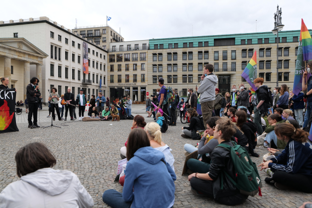 Eine Gruppe von Menschen sitzt auf dem Boden vor einer Menge, die Fahnen und Schilder hält, mit einer Person, die ein Mikrofon, ein Schild und Gebäude im Hintergrund hält, während einer Anti-Schwulen-Demo in Berlin, Deutschland.