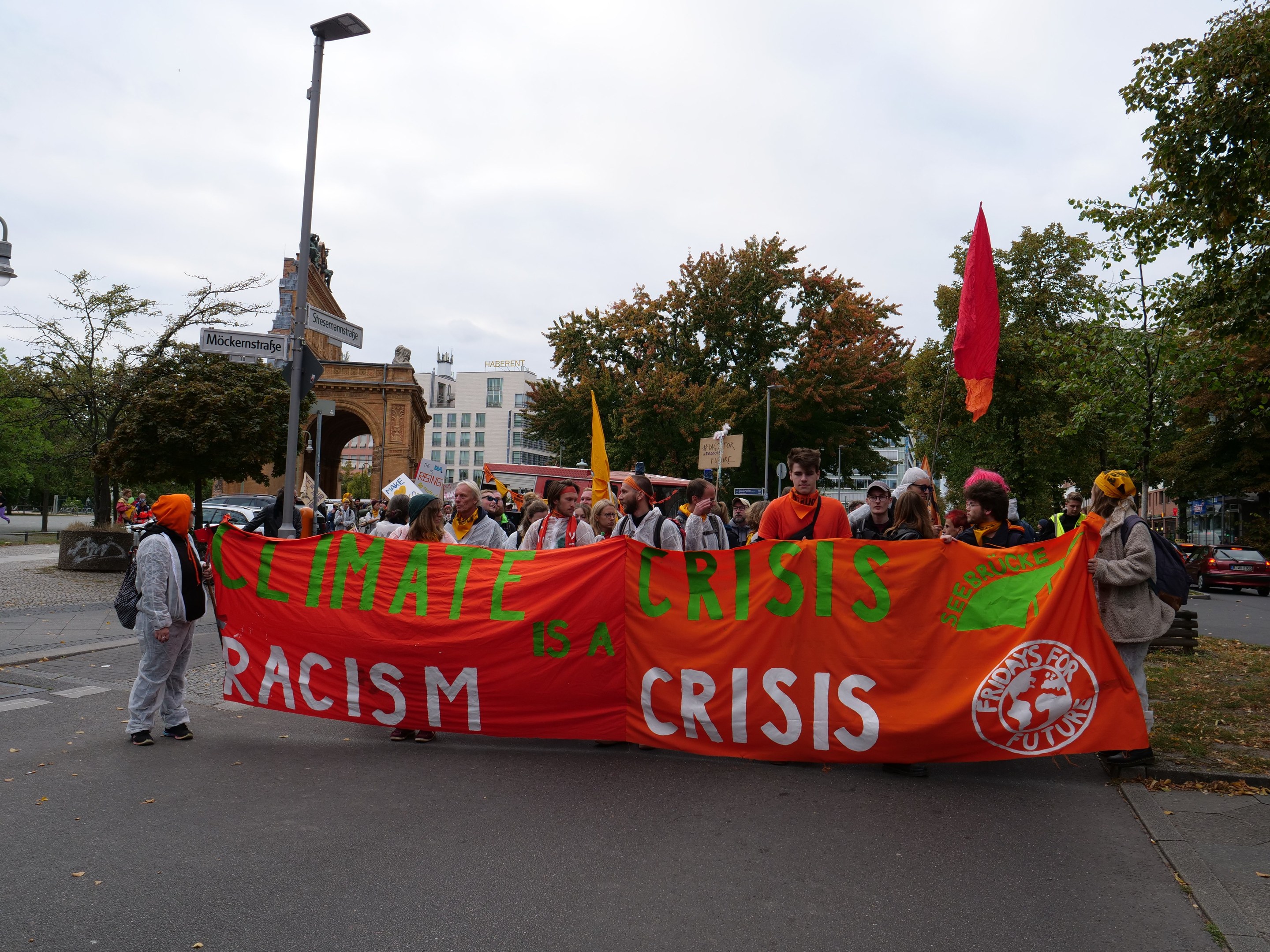 Eine Gruppe von Menschen marschiert auf einer von Bäumen gesäumten Straße und hält ein Banner mit der Aufschrift "Klimakrise ist eine Krise" hoch, mit Fahrzeugen, Gebäuden und einem klaren blauen Himmel im Hintergrund.