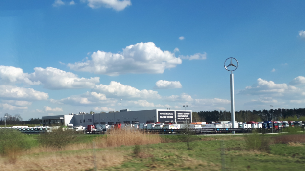 Mercedes-Benz-Fabrik in Deutschland mit Fahrzeugen auf der Straße, umgeben von Gras, Pflanzen, Bäumen, Gebäuden und Strommasten, einschließlich einem mit einem Logo.