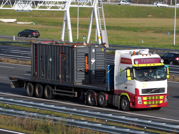 Ein Lkw mit einem großen Behälter fährt auf einer Autobahn mit anderen Fahrzeugen, Masten, Bäumen, Gebäuden und einem klaren blauen Himmel im Hintergrund.