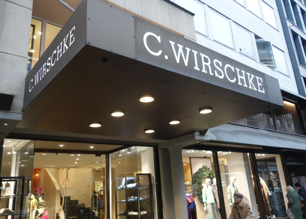 Ein C Wirschke Store in New York City mit Glaswänden und -türen, Schaufensterpuppen in Kleidern, Deckenbeleuchtung und einigen Menschen, die herumlaufen.