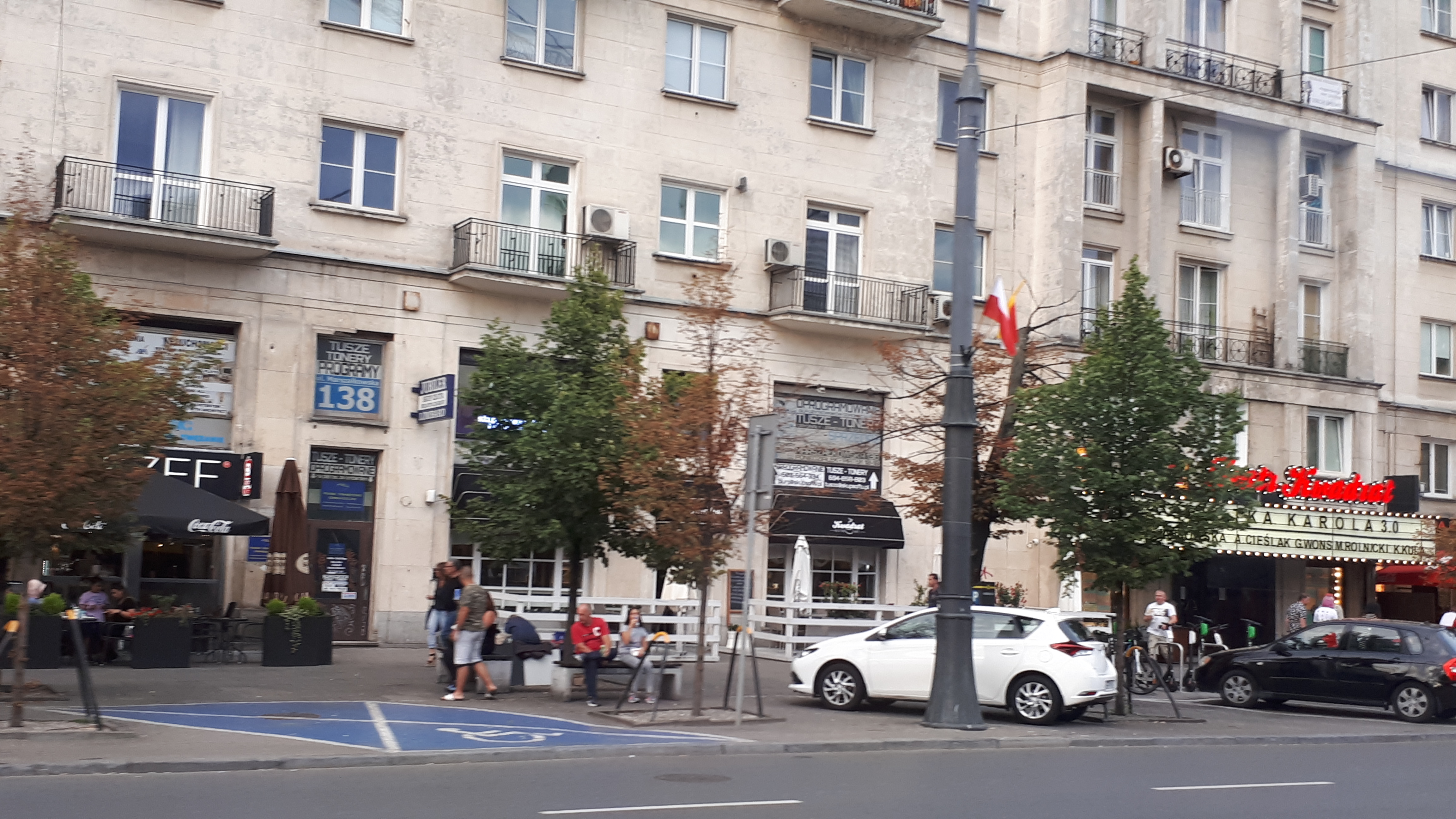 Eine Stadtstraße mit parkenden Autos, gesäumt von Bäumen und einem Gebäude mit Fenstern im Hintergrund, Menschen, die gehen und auf Bänken sitzen, Schilder mit Text und ein Fahnenmast.