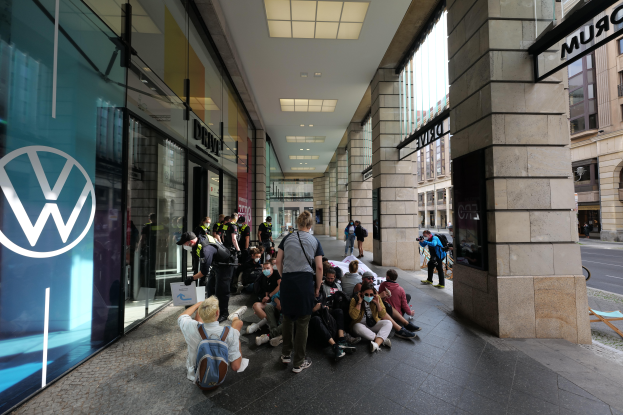 Eine Gruppe von Menschen sitzt und steht vor einem Volkswagen-Geschäft mit Glaswänden und Logo, umgeben von Säulen mit Namensschildern, mit Gebäuden und Deckenlampen im Hintergrund.