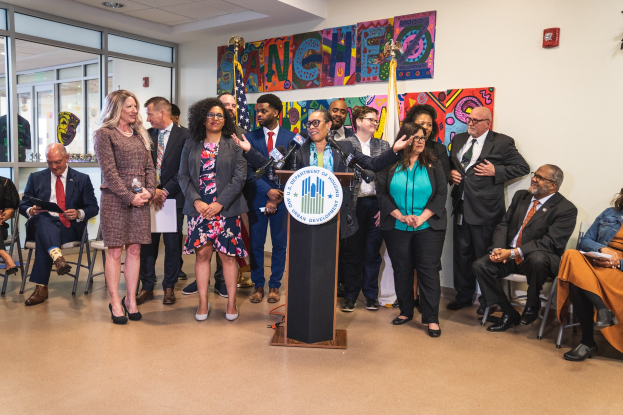 Gruppe von Menschen vor einem Podium mit einem Mikrofon und einer Tafel mit der Aufschrift "Sancho Stadtrat tritt dem Stadtrat bei", mit sitzenden und stehenden Teilnehmern, Flaggen, einem Feuerlöscher und Deckenleuchten.