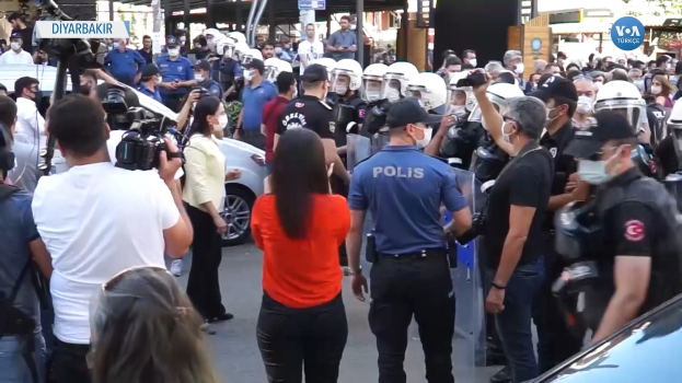 Eine Gruppe von Menschen vor einer Reihe von Helmträgern der Polizei mit Kameras, Fahrzeuge auf der Straße, Gebäude und Bäume im Hintergrund, mit Schildern, die "türkische Polizei im Konflikt mit Demonstranten in Ankara" lesen.