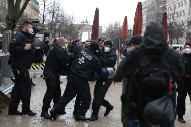 Eine Gruppe von Polizisten in schwarzer Einsatzausrüstung und Masken geht eine Straße in Berlin, Deutschland, entlang, mit Gebäuden, Bäumen, Schirmen, Pfählen, Fahrzeugen und einem sichtbaren Himmel im Hintergrund.
