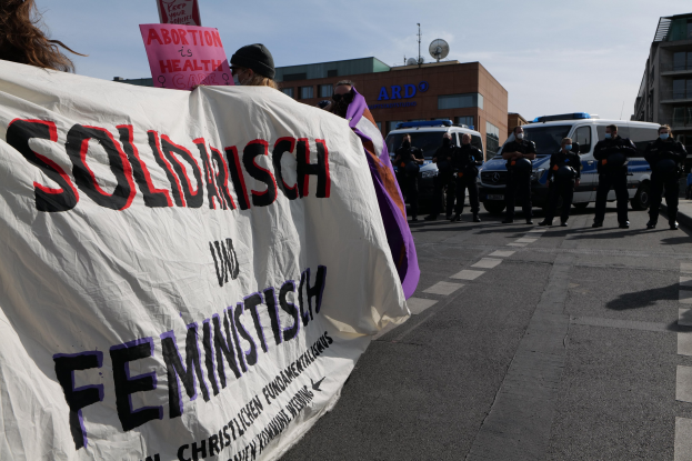 Eine Gruppe von Menschen, die eine 'Solidarität und Feminismus'-Fahne schwenken, geht eine Straße entlang mit geparkten Fahrzeugen, Gebäuden, einer Schüsselantenne und einem klaren blauen Himmel im Hintergrund.