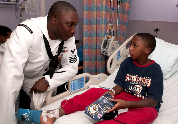 Ein junger Junge sitzt in einem Krankenhausbett und hält ein Buch in der Hand, während ein Mann in weißer Uniform neben ihm steht und von medizinischen Geräten, einem Vorhang und einer Wand umgeben ist.