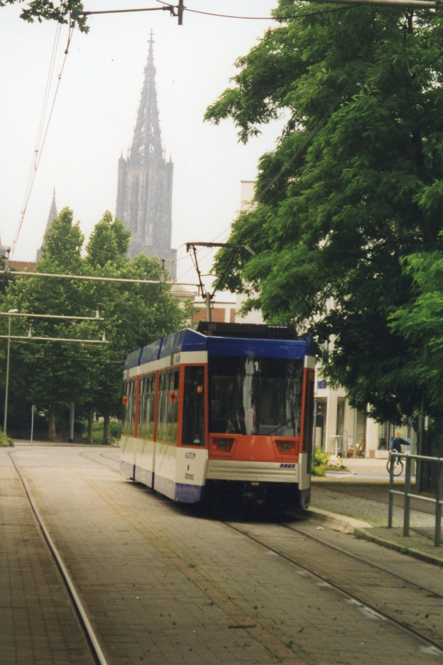 Roter und weißer Zug fährt auf Schienen neben einem hohen Gebäude, mit einer Person, die auf einem Fußweg rechts neben den Schienen Fahrrad fährt, Bäume säumen die Schienen und im Hintergrund sind Gebäude und ein klarer blauer Himmel zu sehen.
