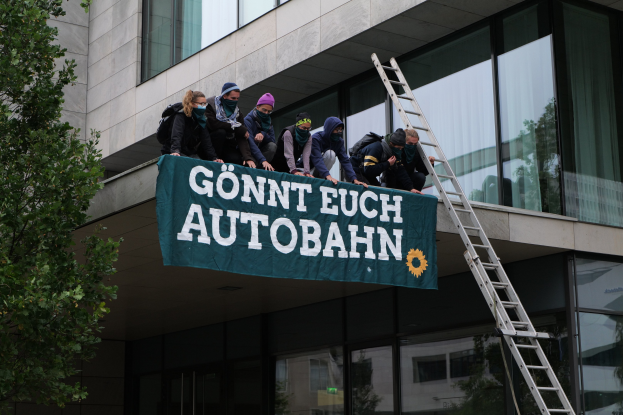Gruppe von Menschen auf einem Gebäudedach mit einem Banner, einem Baum links und Glasfenstern im Hintergrund, wahrscheinlich im Rahmen einer Protestaktion für die deutsche Regierung.
