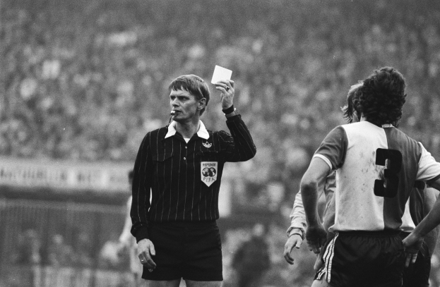 Schwarzes und weißes Bild eines Schiedsrichters mit roter Karte vor einer Menge in Fußballuniformen, mit einem Zaun im Hintergrund.