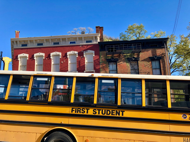 Gelber Schulbus mit der Aufschrift "First Student" vor einem roten Backsteingebäude geparkt, mit ein paar sichtbaren Passagieren, Bäumen und einem klaren blauen Himmel im Hintergrund.