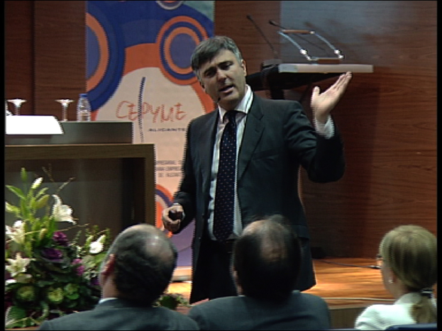 Ein Mann in einem schwarzen Blazer steht an einem Podium mit Mikrofonen, spricht in ein tragbares Mikrofon, mit einer Blumenanordnung und einem Tisch mit einem Buch und einer Wasserflasche in der Nähe, während drei Personen auf Stühlen vor ihm sitzen, vor einer braunen Wand und einer bunten Tafel mit Text.