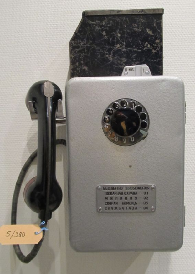 Ein altmodisches schwarzes Münztelefon mit einem weißen Hörer hängt an einer Wand, ein Schild mit Text daran befestigt.