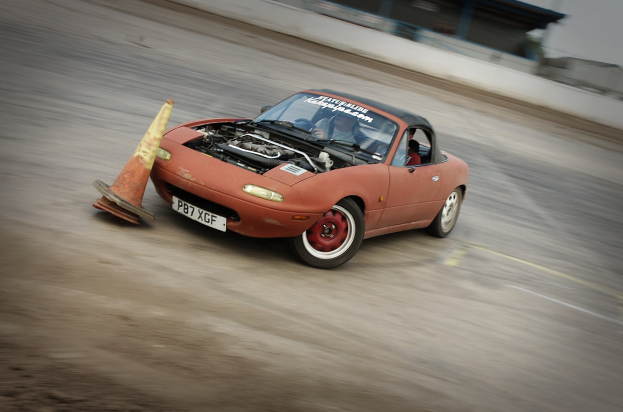 Eine Person fährt ein rotes Auto auf einer Rennstrecke mit einem Verkehrskegel vor sich, Hintergrund unscharf, was auf hohe Geschwindigkeit hindeutet.