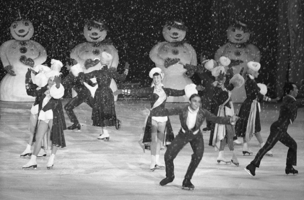 Eine Gruppe von Menschen in Kostümen auf einer Eisbahn im Schnee, mit Schneemännchen im Hintergrund, in Schwarz-Weiß aufgenommen.
