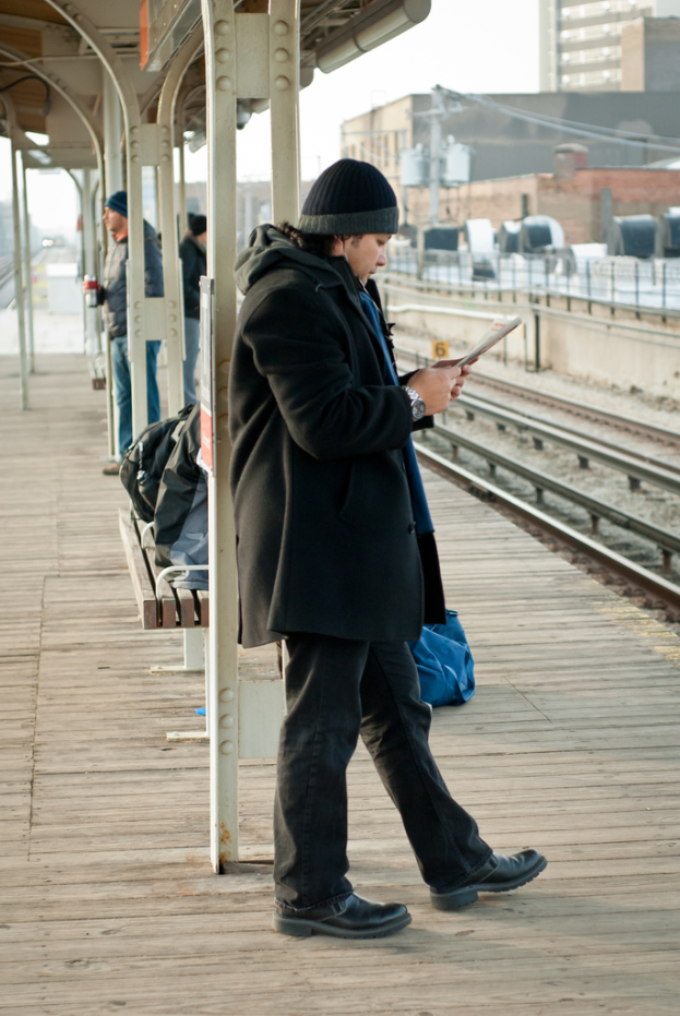 Ein Mann in einer schwarzen Jacke steht an einem Bahnhof, mit einem Eisenpfahl und einem Schuppen im Hintergrund und Bahnschienen und braunen Gebäuden rechts.