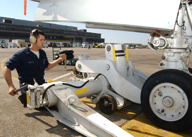 Ein Mann mit Kopfhörern steht neben einem Flugzeug auf einer Landebahn, mit Fahrzeugen, Gebäuden und einem klaren blauen Himmel im Hintergrund.