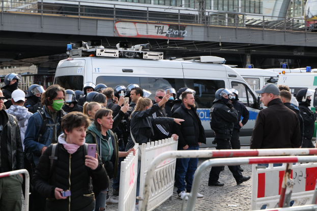 Eine Gruppe von Menschen steht in der Nähe von Polizeiwagen mit Barrieren im Vordergrund und einer Brücke im Hintergrund während einer Demonstration in Berlin, Deutschland.